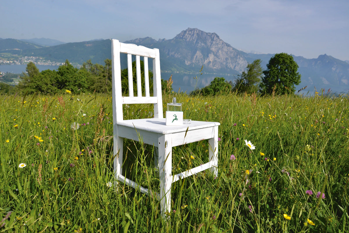 Blick vom Gmundnerberg auf den Traunstein. Auf einer Blumenwiese steht ein weißer Sessel mit einem Flakon GRÜNER HIRSCH.